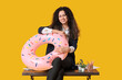 © Pixel-Shot - Happy female office worker with inflatable ring sitting on table against yellow background. Summer vacation concept