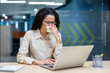 © Liubomir - Woman in office setting working on laptop, holding tissue, showing signs of being unwell. She wears glasses and focuses on her work, reflecting concept of managing health in professional environment.