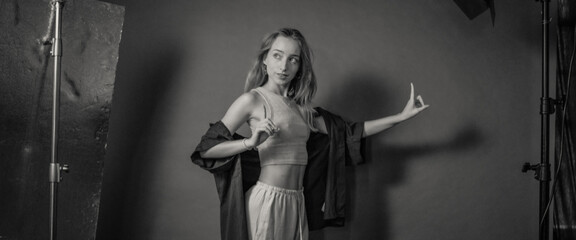 A young woman artistically poses with expressive gestures, dressed in a stylish casual outfit, standing in a studio setting with professional lighting equipment.