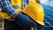 © rookielion - A worker wearing gloves holds a yellow safety helmet next to a solar panel, emphasizing safety in renewable energy