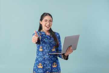 Government worker woman in blue batik uniform holding laptop while doing thumb up gesture.