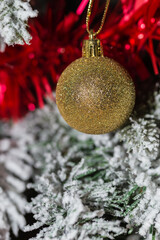 Vertical shot of a shiny golden ball hanging on a green and snowy Christmas tree decorated with red tinsel. Christmas ornament. Wallpaper. Copy space. Christmas concept, festivity and decoration