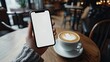 © KYR - Woman's hand holding a smartphone with a white screen, a cup of coffee beside it on a trendy table in a stylish cafe