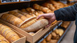 © OMD - Hand picking up a freshly baked loaf of bread from a wooden tray at a bakery or grocery store, with several loaves of bread displayed on shelves.
