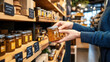 © Natalia - Person holding a jar of honey in a specialty food store. Shelves are stocked with various jars of honey and other products, with chalkboard labels.