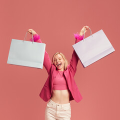 Happy, smiling, joyful young woman in pink jacket raising two shopping bags against pink studio background. Giant sale season. Concept of Black Friday, Cyber Monday, shopping, sales