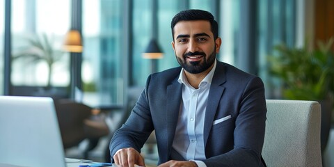 Smiling businessman sits at a desk with a laptop.