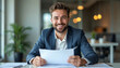 © Ali Hamza Tullah - Portrait of a smiling and successful young businessman sitting in the office at the table, holding documents and papers for a deal in his hands, confidently looking at the camera