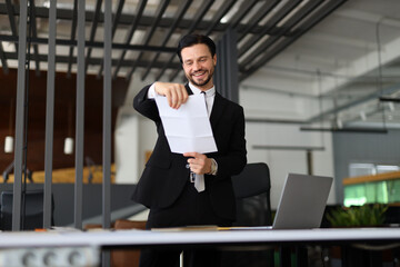 A man in a suit is holding a piece of paper and smiling. He is standing in front of a desk with a laptop and a chair. Concept of professionalism and confidence