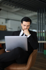 Wall Mural - A man in a suit is sitting in a chair with a laptop open in front of him. He is deep in thought, possibly working on a project or problem. Concept of focus and concentration