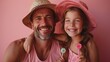 © amixstudio - A joyful father and daughter pose together, both wearing pink hats and smiling broadly. The daughter holds colorful lollipops, and they are set against a pink background