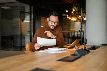 Wall Mural - serious male accountant in glasses examining spreadsheets and reports while working at desk in offic