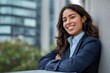 © Viktor - Young hispanic business woman in professional attire smiles confidently leaning on a city wall. Modern executive woman with curly hair and attractive features.