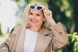 © deagreez - Portrait of positive happy retired woman smile spring good mood take off sunglass weekend wear beige coat outdoors outside park street