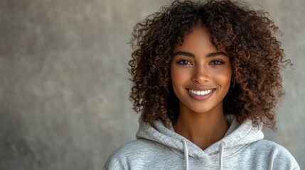 A young African American woman with a radiant smile poses confidently in a casual gray hoodie against a textured gray background, exuding modern urban style.