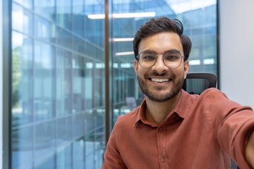 Wall Mural - Confident young businessman smiling for selfie using smartphone in modern office environment. Displays success, positivity, and professionalism. Perfect for promoting positive workplace culture