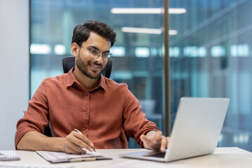 young professional sitting at desk in modern office using laptop and taking notes. man is focused on