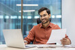 © Liubomir - Young professional man wearing glasses working happily at laptop in modern office. Holding document, showcasing productivity and success. Captures business environment, with sense of achievement