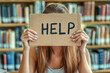 © CozyDigital - Help. Young girl sits in library with cardboard sign that says 'Help.'