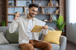 © Tetiana - Joyful man celebrating with fist pump while holding important letter in cozy living room setting