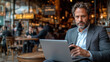 © aekkorn - Phone Call : A man in a suit sits in a cafe, working on his laptop and using a cell phone. amidst a bustling atmosphere, surrounded by technology and people.