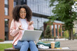© Liubomir - Joyful African American female student engaging in online learning and video call on laptop outdoors. Relaxed and focused on communication and education with headphones and phone nearby.