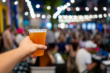 Wall Mural - Hand holding a plastic cup of beer with a blurred festival background. Bokeh lights create a lively atmosphere, capturing the essence of a fun outdoor event. Perfect for themes of celebration