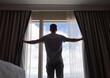 © kieferpix - Young man looking out window in the morning opening curtains. Concept of a new day and joyful weekend.