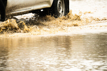 A car is driving through a flooded street with water splashing up. The car is covered in mud and the water is murky. The scene is chaotic and dangerous, as the driver must navigate through the water