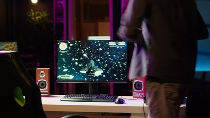 Wall Mural - Man playing SF space videogame on PC screen in neon illuminated apartment living room. Gamer enjoying spaceship flying game on computer desktop display in RGB lit home late at night, camera A