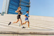 © muse studio - A fit couple running up urban stairs, with modern architecture towering behind, under a clear blue sky.