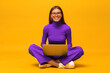 © Damir Khabirov - Happy millennial girl in green sitting on floor with laptop on knees on yellow background