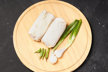 Two frozen lumpia filled with bamboo shoots and meat, with white wrappers, ready to fry. Served with green chili peppers and scallions, photographed on a round wooden tray against a dark background.