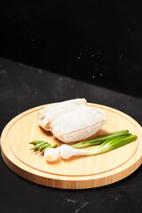 Two frozen lumpia filled with bamboo shoots and meat, with white wrappers, ready to fry. Served with green chili peppers and scallions, photographed on a round wooden tray against a dark background.