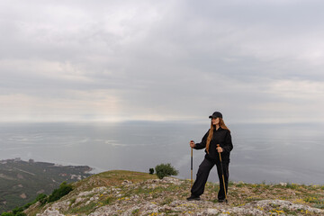 Wall Mural - A woman stands on a hill overlooking the ocean, holding two walking sticks