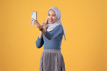 Wall Mural - happy asian indonesian muslim woman holding smart phone giving victory finger gesture on isolated yellow background