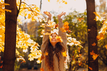 Smiling young woman with hands up throws up the autumn leaves in the forest at sunset