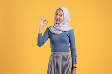 Wall Mural - happy asian indonesian muslim woman giving ok finger gesture on isolated yellow background