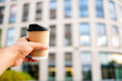 © pavel siamionov - A hand holding a disposable coffee cup with a black lid, against a blurred urban background. The focus is on the cup, highlighting the concept of takeaway coffee in a city setting