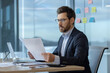 © Liubomir - Businessman in office wearing suit and glasses, focused on analyzing documents at desk. Laptop and smartphone present, creating professional atmosphere.