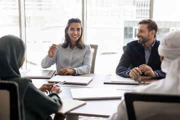 Wall Mural - Happy beautiful Hispanic businesswoman talking to Arab partners in Muslim clothes, sitting at meeting table, speaking, smiling, laughing, discussing creative ideas for collaboration