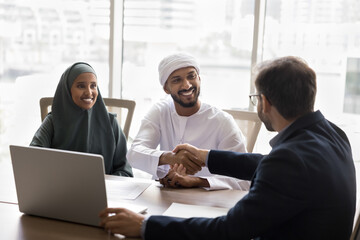 European business professional selling property to young Arabian couple, shaking hands with happy husband in Muslim clothes, giving consultation about real estate investment, mortgage