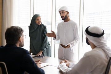 Wall Mural - Couple of young Arabian managers in Muslim clothes presenting project to European business partners, speaking to colleagues sitting at meeting table, discussing company selling, investment