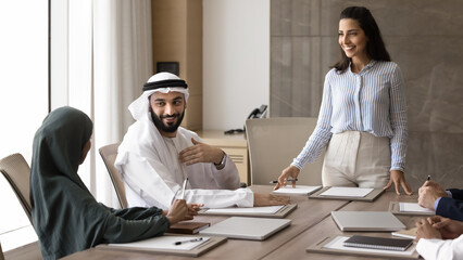 Wall Mural - Happy successful young Hispanic business coach woman training Arabian professional men and women. European female speaker holding meeting with group of Muslim entrepreneurs at large table. Banner