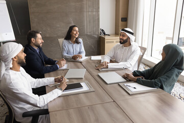 Wall Mural - Positive diverse business team of Arabian and European entrepreneurs meeting for brainstorming, planning company management, talking, laughing. Multiethnic board of directors high angle shot