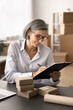 © fizkes - Middle-aged female small business owner sit at desk writes notes on clipboard, surrounded by cardboard boxes, involved in logistics, order management workflow. Preparing shipment or handling inventory
