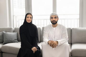 Handsome bearded Arabic husband in Muslim clothes and positive young wife with open face sitting on comfortable couch, looking at camera, posing for Arabian couple home portrait