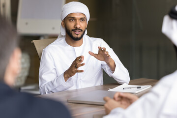 Wall Mural - Positive engaged young Arabian business professional man in white Muslim clothes explaining creative ideas for project to colleagues at meeting, brainstorming on strategy, speaking