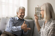 © fizkes - Older hoary husband sit on sofa at home communicates with mature wife using gestures. Older man with hearing loss, physical disability interact with smiling positive spouse showing each other symbols