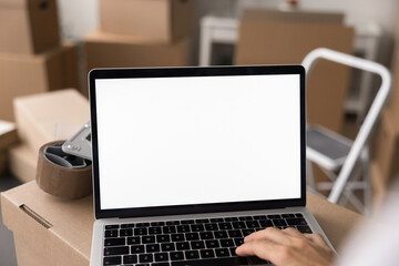 Female working on laptop with white mock up in stockroom filled with stacked boxes. Electronic commerce worker managing inventory, processing orders, labeling packages, coordinate shipments, close up